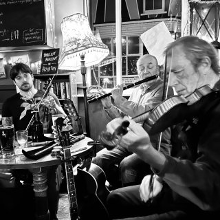 A man playing fiddle and a man playing flute while another fiddler looks on, all of them gathered around a pub table.