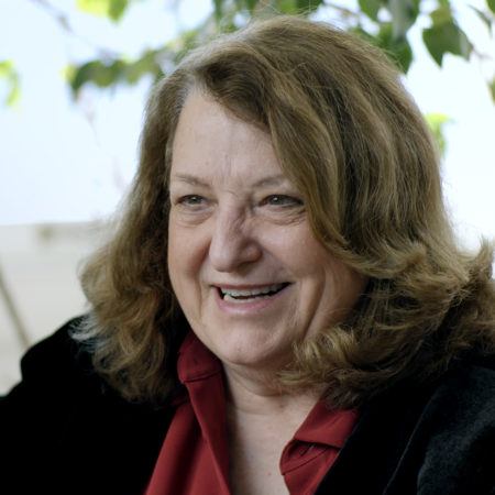 Lynn Hershman Leeson in a red shirt black blazer in front of plants.