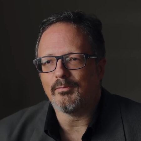 Rafael Lozano-Hemmer with glasses and a beard wearing a dark suit jacket sits against a dark background.