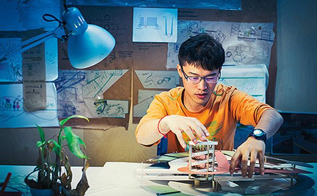 Male student at desk working with design model, with schematics posted behind him