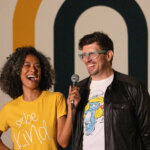 A woman of color in a bright yellow t-shirt that says Be Kind on the front smiles and holds a mic toward the man next to her. The man is white and wears thick rimmed glasses, a white t-shirt, and black leather jacket. He is also smiling.