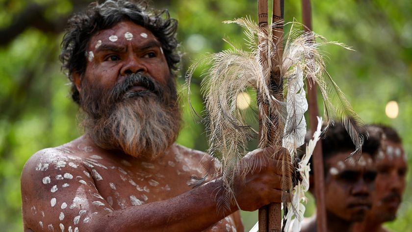 An Indigenous Australian man in traditional dress holding a wooden weapon with feathers.