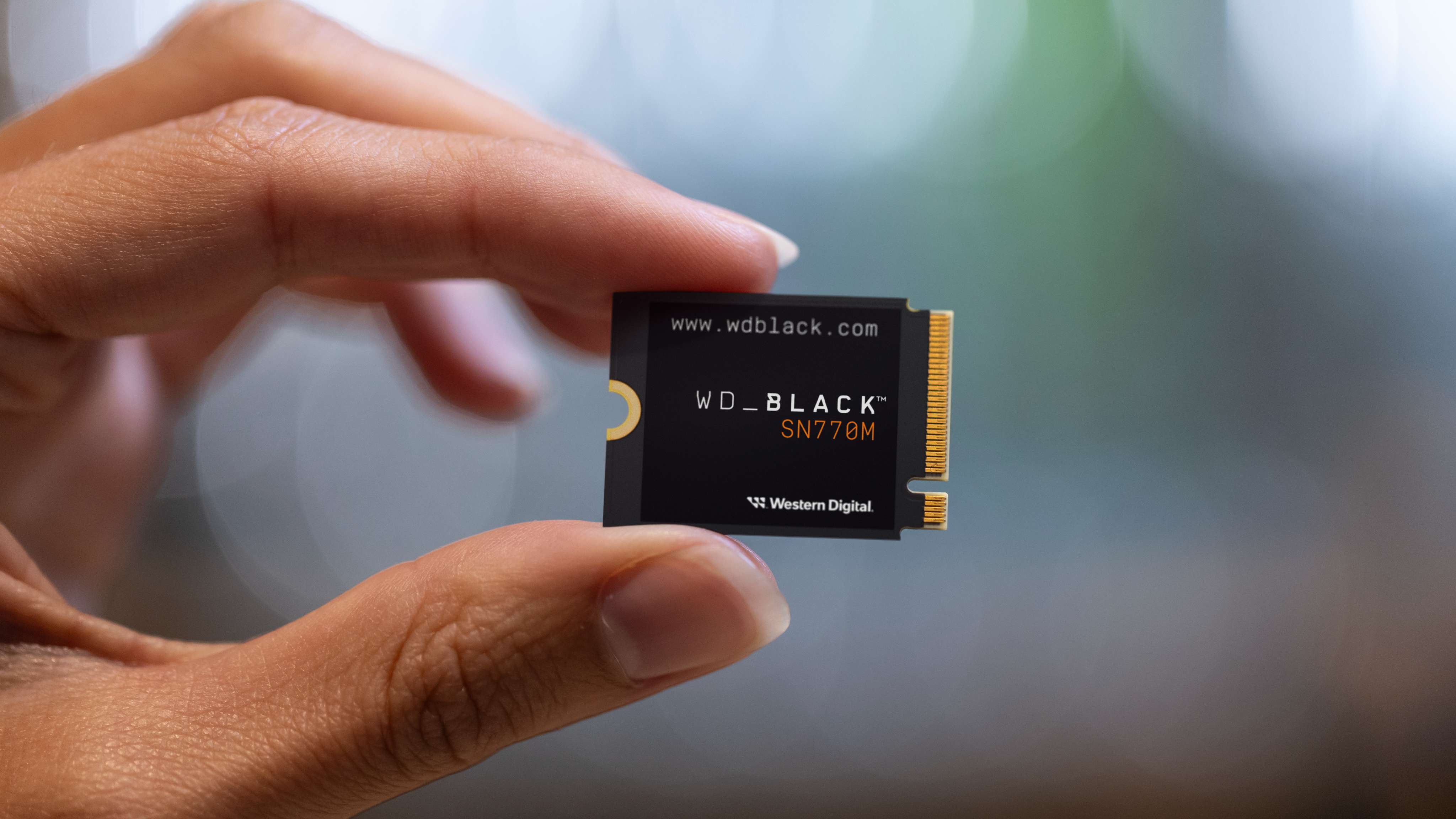 A hand delicately holds a WD_BLACK SN770M SSD against a blurred background. The focus is on the compact, rectangular storage device, conveying precision and technology.