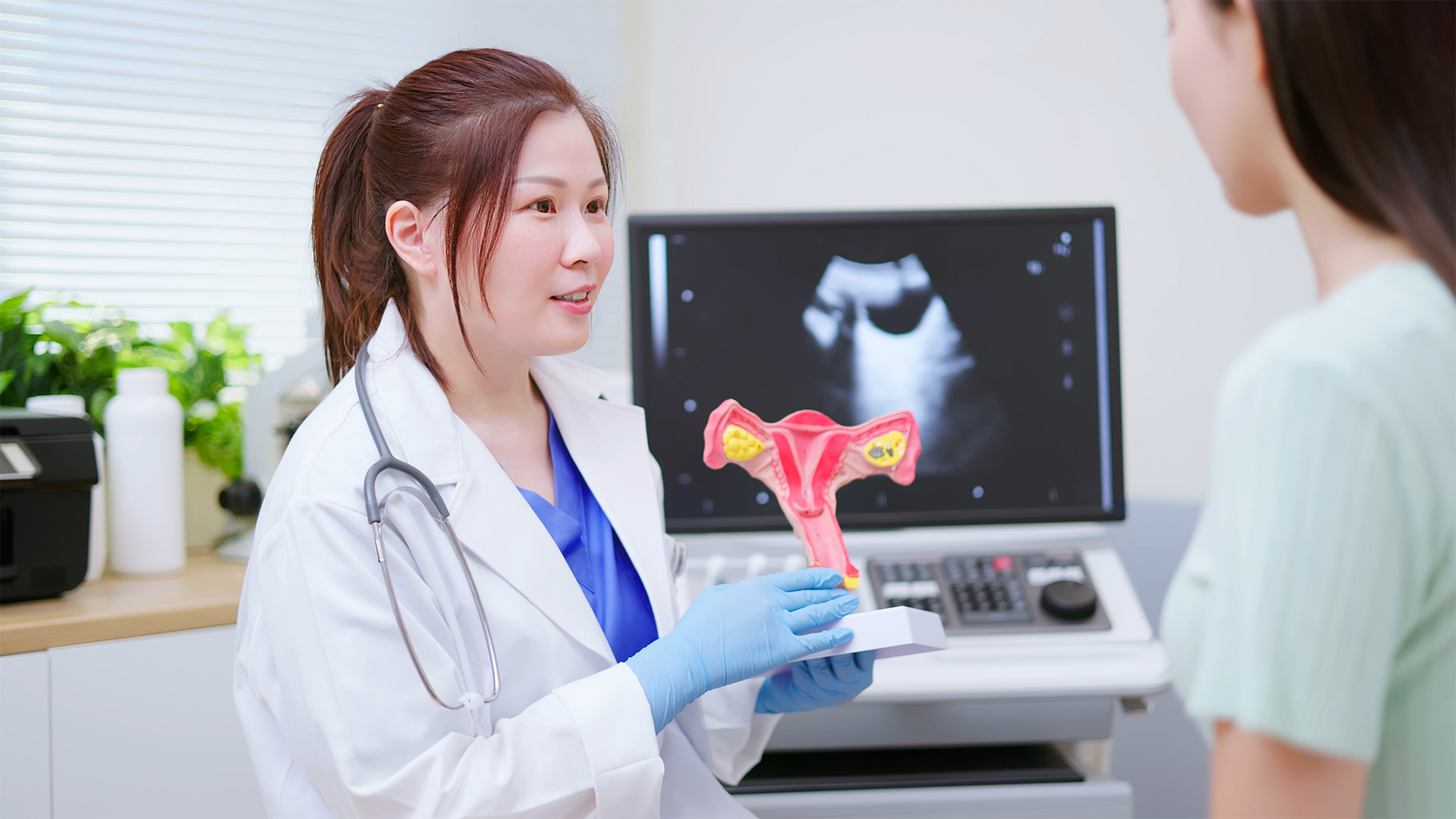An Asian woman with brown hair in a pony tail wearing a white lab coat and blue gloves holds up a model of the female reproductive system.
