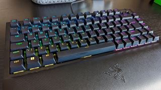 A black gaming keyboard with RGB backlit keys on a desk. Keys display a gradient of colors, creating a vibrant and dynamic effect. Razer logo visible.