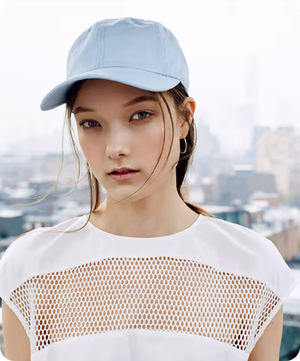 Young woman wearing a light blue cap and white top with mesh detail, standing outdoors with a cityscape background.