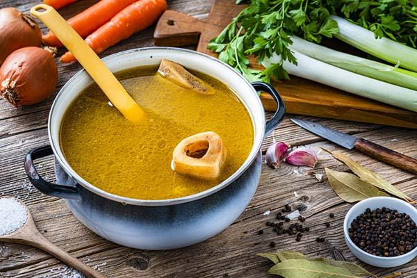 A pot of bone broth sits on a rustic wood table, with a ladle and pieces of bone in the broth; around the pot are vegetables: onions, carrots, and celery stalks, along with a few cloves of garlic and a small bowl of capers.