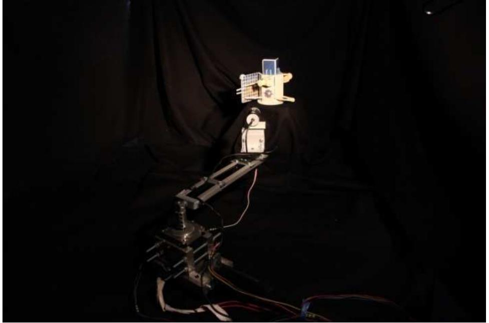 Figure 11: The set-up with the fixed scaled model of the deputy and the camera representing the chief spacecraft at the tip of the arms. Black curtains help in reproducing lighting conditions in space.  eccentric, parking orbit in which the chief maintains a safe distance before rendezvous begins). The chief (i.e. the camera) can capture the images of the deputy and, by means of the computer labelled as chief’s OBDH, evaluate the relative position and attitude and define the requested control actions, which are then transmitted to the arm’s computer to be  implemented [14]. 