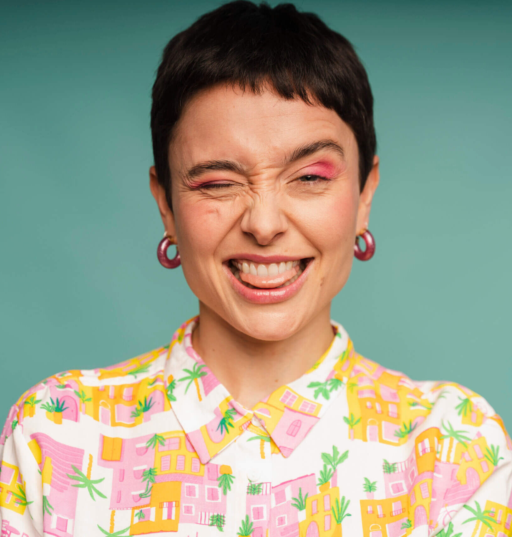 Eine Person mit kurzen dunklen Haaren steht vor einem türkisfarbenen Hintergrund. Sie zwinkert, lacht breit und streckt spielerisch die Zunge heraus. Sie trägt ein buntes Hemd mit einem Muster aus Häusern in Pink- und Gelbtönen sowie Palmen. In den Ohren hat sie große pinke Creolen.