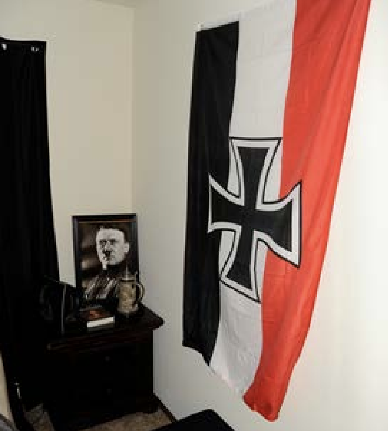 A Nazi battle flag flies on the wall next to a nightstand, on which a picture of Adolf Hitler is visible.