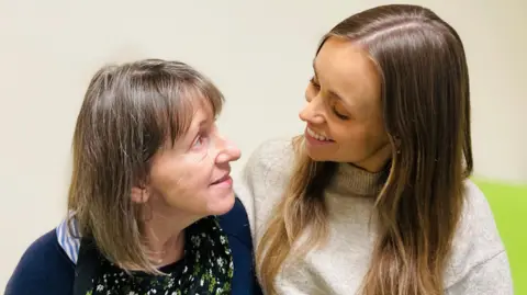 Patient Samantha Denison is sitting on a hospital bed and looking at her daughter Steph. Samantha has shoulder length brown hair and wears a dark blue cardigan over a floral top. Steph has long brown hair and wears a light grey,  high-neck jumper.