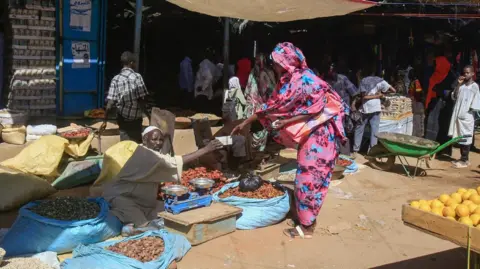 A woman wearing a patterned pink and blue dress buys from a market trader, who is sat on the floor at Abu Shouk's market, surrounded by produce.