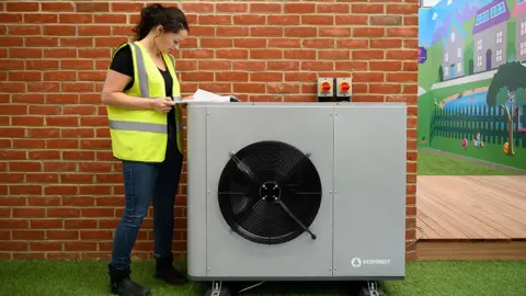 Two men install a heat pump outside a house. Both are crouching either side of the heat pump. The man on the left is wearing a green t-shirt and shorts. The man on the right is wearing a black hoodie, trousers and a baseball cap.