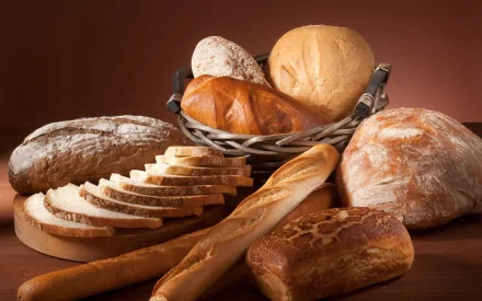 HD desktop wallpaper featuring a variety of fresh bread, including sliced loaf, baguette, and round buns arranged on a warm brown background.