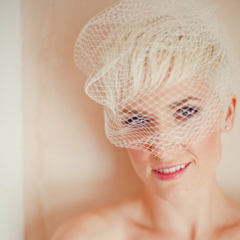 Smiling bride peers at camera from behind a vintage birdcage veil. 