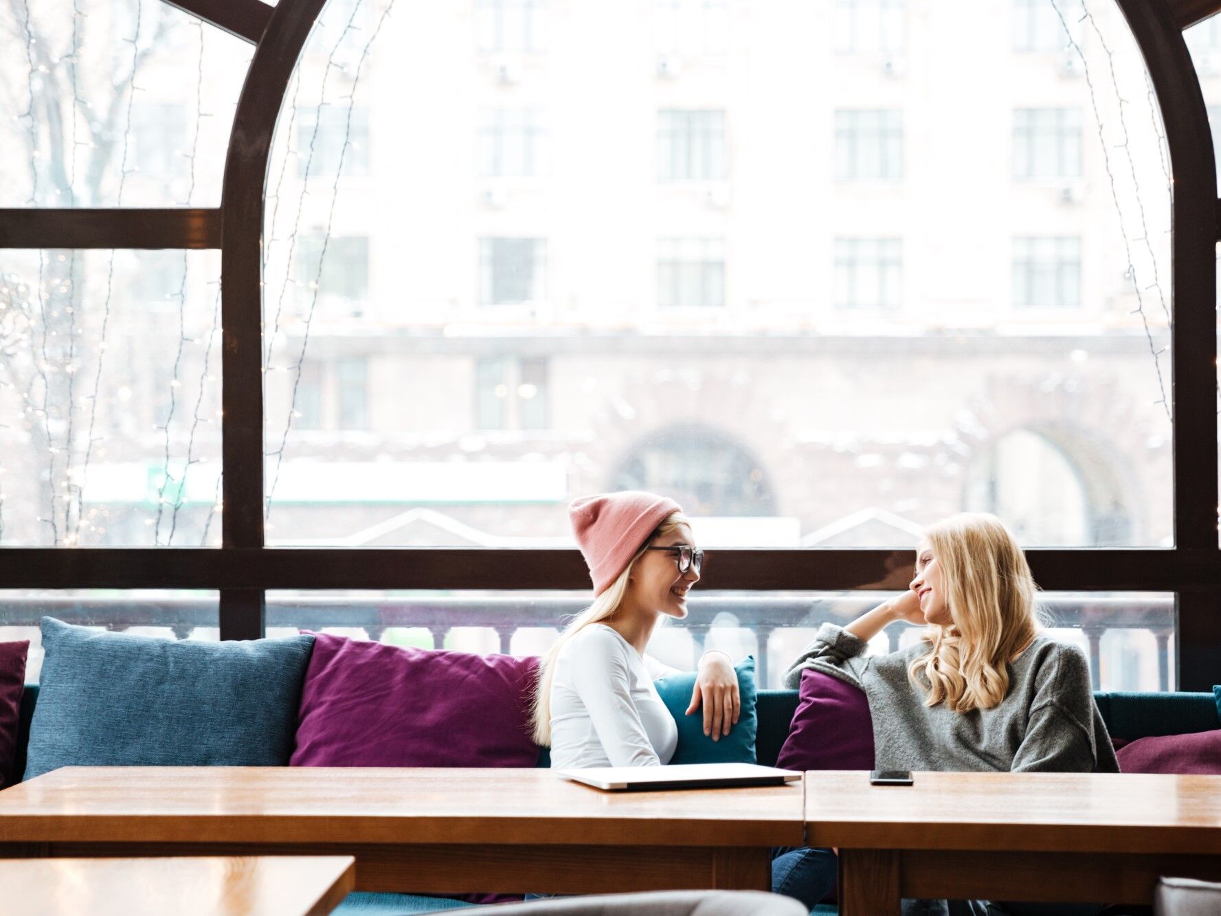 Questions to Ask Your Girlfriend - Two women chat while sitting on a couch in front of a large window. 