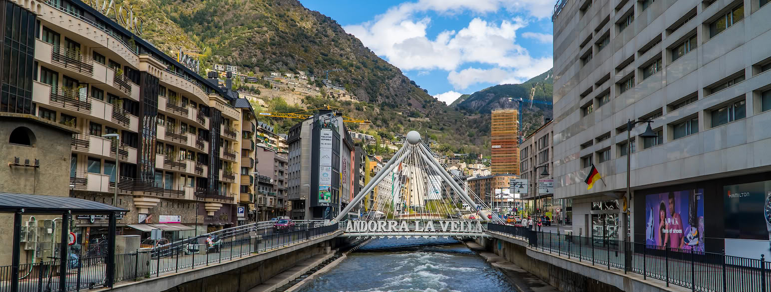 En elv renner under en bro mellom høye bygninger i en by. På broen står det ANDORRA LA VELLA. I bakgrunnen er det bratte fjell og blå himmel med noen skyer. Foto