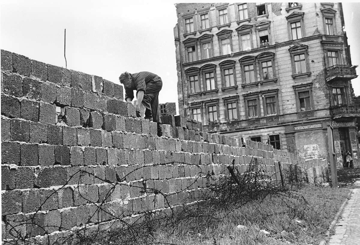 En mann bygger en mur ved siden av en bygård. På bakken ligger piggtråd. Foto