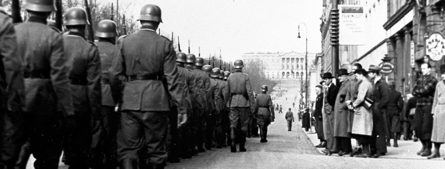 Svart-hvitt foto Karl Johans gate i Oslo på vei opp mot slottet marsjerer tyske soldater med uniform og hjelm. På fortauet står folk og ser på. 