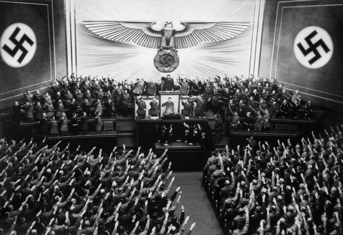 Svart-hvitt foto av en stor sal med store hakekors og den tyske ørnen på veggen. Hitler står på et podium omringet av flere andre. Nede i salen sitter det tett i tett med folk. Alle hever armen i nazihilsen mot Hitler. 