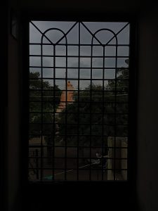 A view from inside a dimly lit room through a barred window, showing an orange temple spire amidst trees against a cloudy sky.
