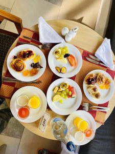 A round wooden table set for a meal with several plates containing pastries, jams, fresh fruits, and small glasses of different fruit juices. There are napkins, utensils, a salt and pepper shaker, and a bottle of water. The setting is bright, with natural light illuminating the dishes