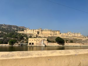 Looking at Amber Fort across the water (Front View). It’s a fortress/palace with many walls and turrets built on a hill.
