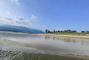 A serene riverside landscape with clear blue skies, scattered clouds, and a distant view of green hills and mountains.
