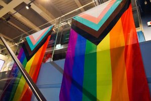 Two Progress Pride flags are hanging on a glass balcony inside a modern building with overhead lighting and exposed beams.