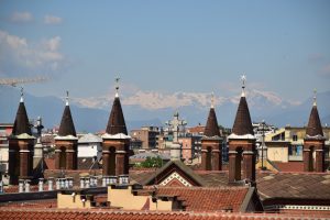 Multiple pointed brick towers topped with star ornaments, with a cityscape and snow-capped mountains in the background under a clear blue sky.