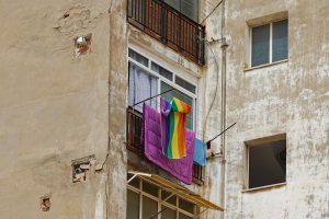 An exterior view of a weathered building wall with visible cracks and patches. A window with a balcony displays a colorful rainbow-striped towel, a purple blanket, and a blue cloth hung out to dry.