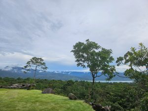 A scenic landscape featuring lush greenery with trees in the foreground and a distant view of mountains partially covered by clouds. The sky is mostly overcast with some patches of blue. 
