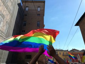 Hands hold up a large rainbow flag in a bright, sunny street surrounded by buildings. Other rainbow flags can be seen in the background, and the clear blue sky contrasts with the vibrant colors of the flags.