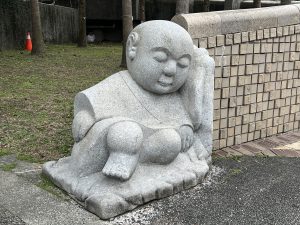 A stone Buddha statue sleeps against a stone wall with trees and grass behind it.
