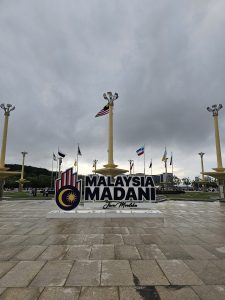 A large sign reading "Malaysia Madani" with a logo featuring the sun and crescent is prominently displayed on a paved area in front of the Prime Minister's Office, Kuala Lumpur Malaysia. Several flagpoles with international flags surround the area.  