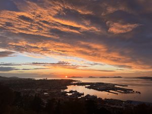 A stunning sunset over Islas Cies, Vigo, with vibrant orange and pink clouds filling the sky. The sun is setting on the horizon, illuminating the harbor below with warm tones. 