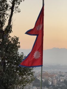 The national flag of Nepal waving on a flagpole with an urban landscape and hazy mountains in the background during sunset