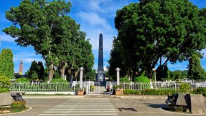 The image shows a scenic park featuring a tall monument in the center, surrounded by lush green trees.