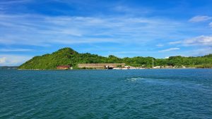 A scenic view of a lush green hill rising from the blue waters, with a small, red-roofed building at the foreground. In the background, a coastline dotted with homes and boats is visible, under a partly cloudy blue sky.