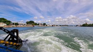 View from the back of a moving boat showing foamy water trail, looking toward a coastal town with various buildings and trees under a partly cloudy sky.