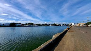 Harbor scene showing calm water, dock, and a small town with boats and buildings, set under a partly cloudy blue sky.