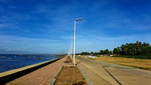 Long coastal walkway lined with modern white streetlights, bordered by a calm sea on one side and grassy land with palm trees on the other.