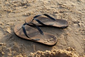 A pair of flip-flops resting in the sandy beach, partially covered in fine grains of sand, with sunlight casting a warm glow on the scene.
