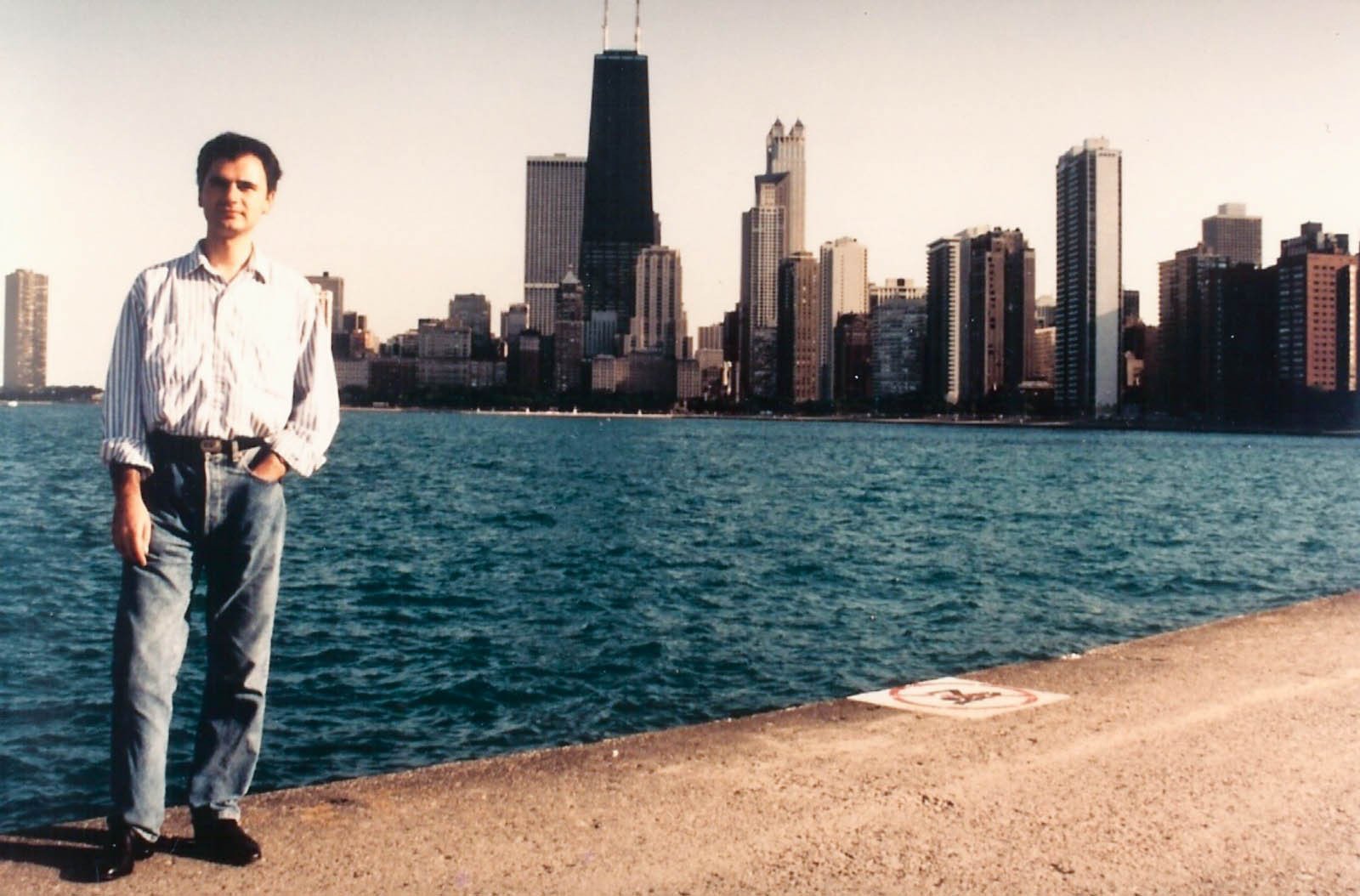 A man stands by the water's edge, overlooking a city skyline with tall buildings, embodying the spirit of writer and journalist Dejan Stojanovic.