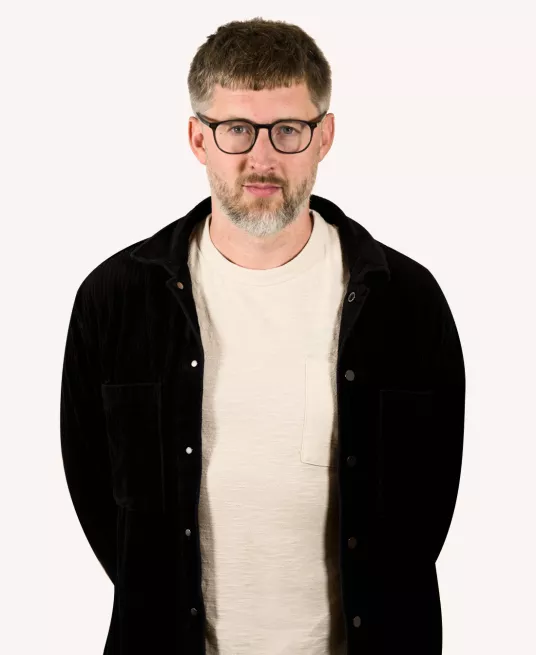 Photo of Fred, a friendly-looking man sporting a salt-and-pepper beard, wearing a white t-shirt and black jacket.