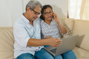 A couple sits together on the couch. The man is holding a computer, and the woman is talking on the phone.