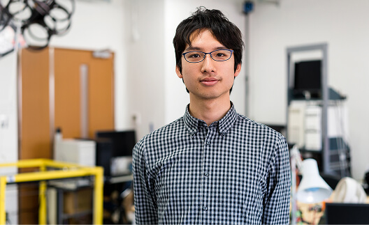 A young man with glasses and a checkered shirt stands in a classroom or lab setting with shelves and equipment in the