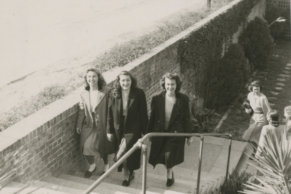 The black and white photograph shows three women walking up a set of outdoor stairs. The stairs have a brick wall on the left side and a metal handrail on the right. The women are wearing mid-20th century style coats and dresses, with one carrying a small purse. They are smiling and walking towards the camera. In the background, more people are visible on the stairs and a garden area is partially visible with neatly trimmed hedges and shrubs.