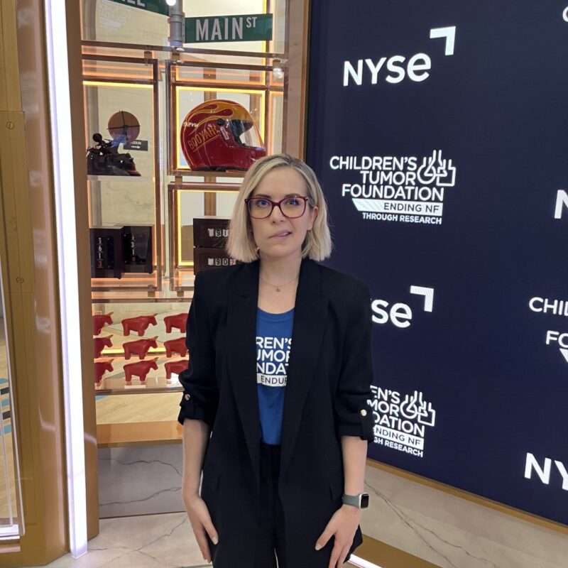 A person stands in front of a display at the NYSE, featuring logos of the Children's Tumor Foundation and street signs for Wall and Main Street.