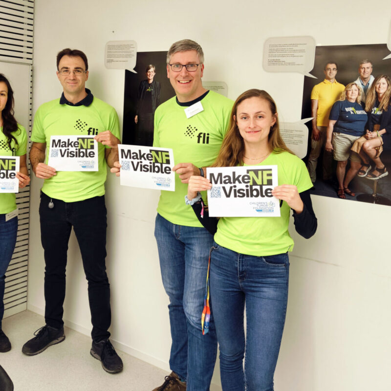 Four people wearing green shirts hold "Make NF Visible" signs, standing in a room with photos and text on the walls.